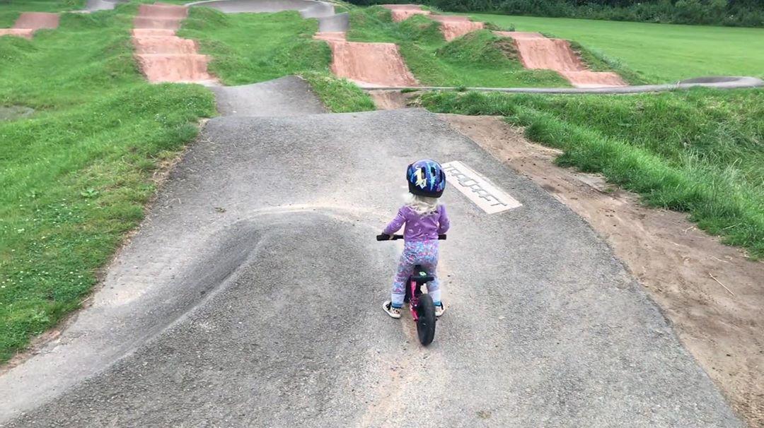 young child sitting on her bike looking out at a large pump track looking like she is about to ride in. taken by instagram @ kidstothefront
